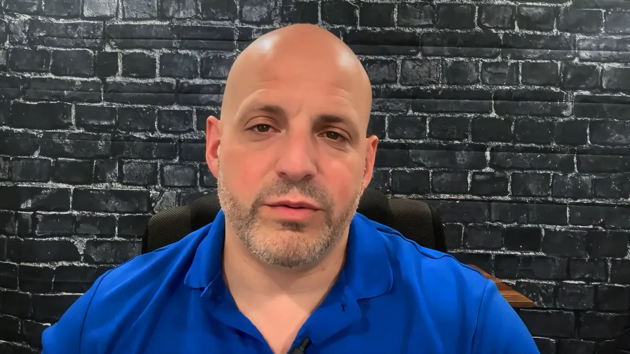 Presenter looking directly at camera against a black brick background, wearing a blue polo — clear, well-lit head-and-shoulders shot