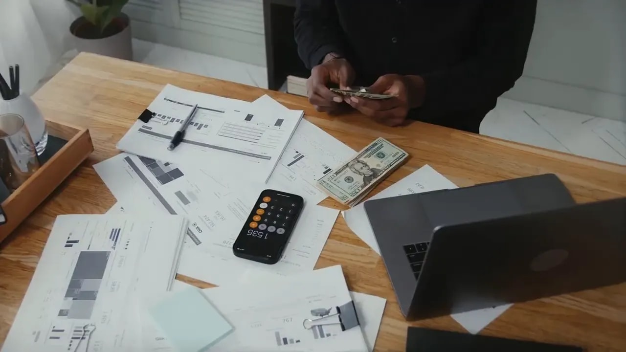 Business documents, cash, calculator, laptop, and phone on a desk