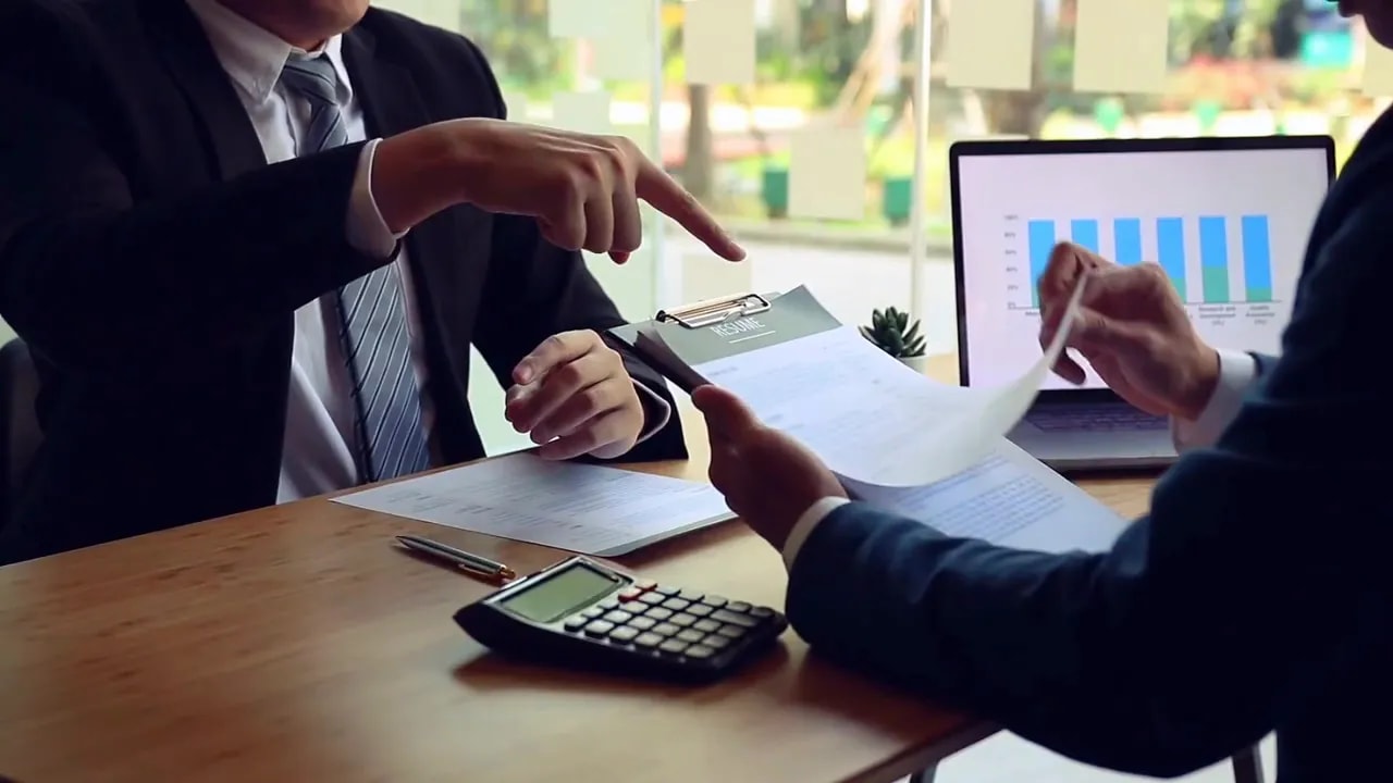 Two professionals reviewing paperwork with a calculator and business chart on a computer monitor during a side hustle or business ownership discussion