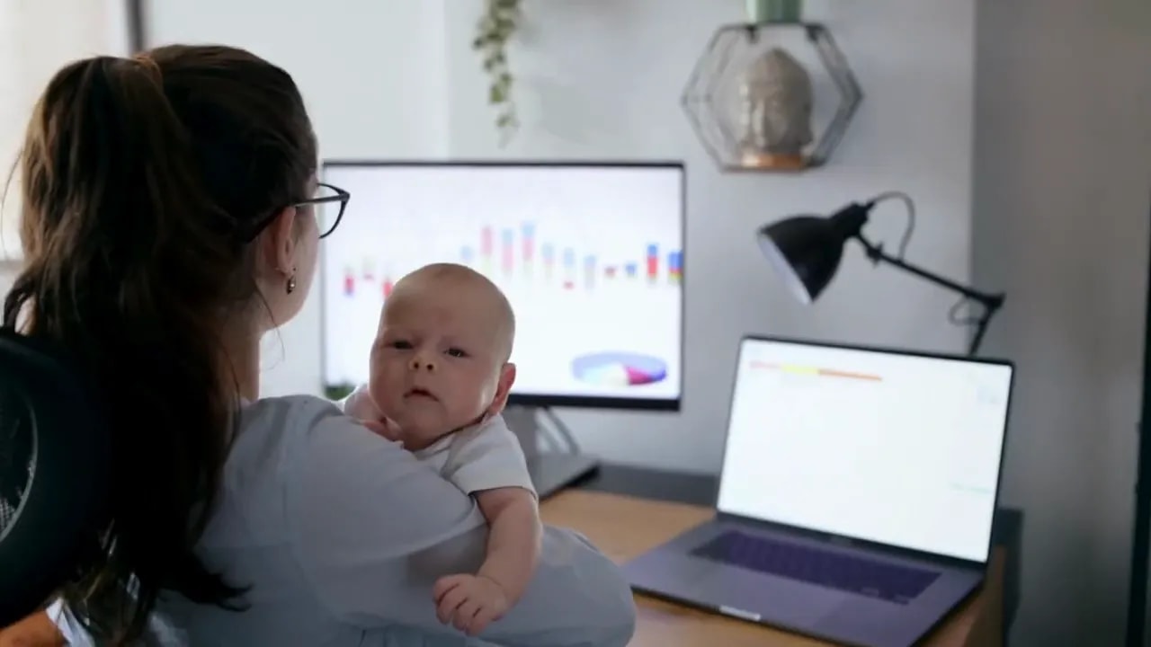 Parent holding a child while working at a desk with computer screens