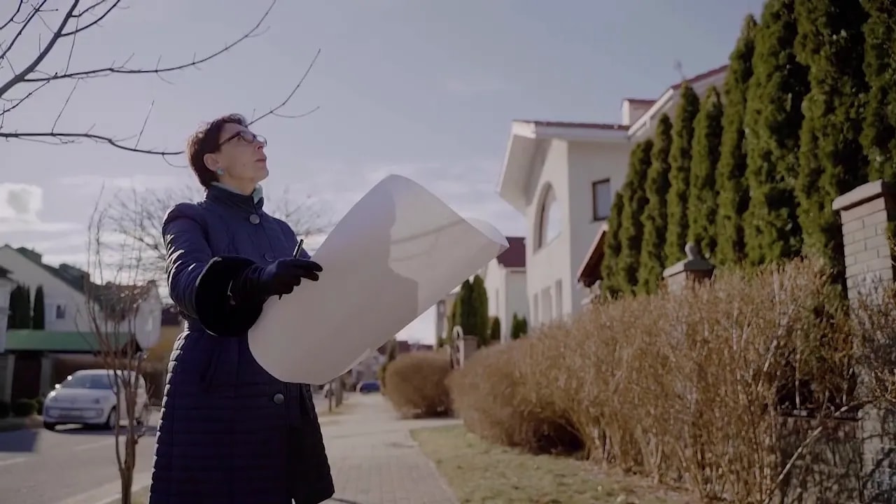 Person outdoors holding property plans and visually inspecting landscaping and hedges beside a residential/commercial property.