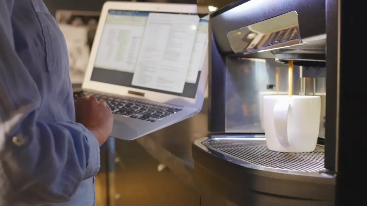 person holding laptop beside office coffee machine pouring into white cup