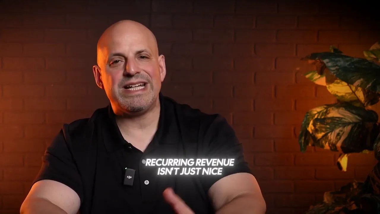 Presenter speaking with clear on-screen text 'RECURRING REVENUE ISN'T JUST NICE' over a warm-lit studio background and plant.